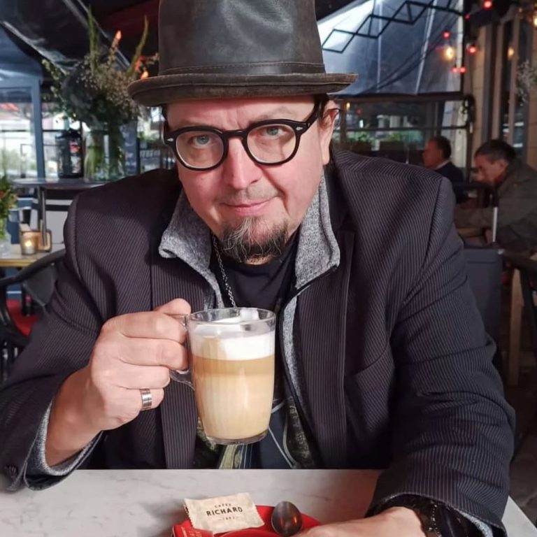 Mann mit Hut und Brille trinkt einen Cappuccino in einem Café. Enrico Palazzo ist ein Musiker, Filmemacher und Schauspieler aus Düsseldorf.