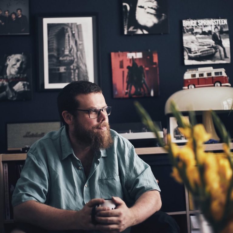 Mann mit Brille sitzt in einem stilvollen Raum, umgeben von Kunstwerken und gelben Blumen.