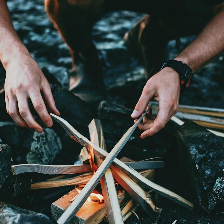 Hände, die Holzscheite um ein kleines Feuer herum anordnen. Survivalcamp, Überlebenstraining.