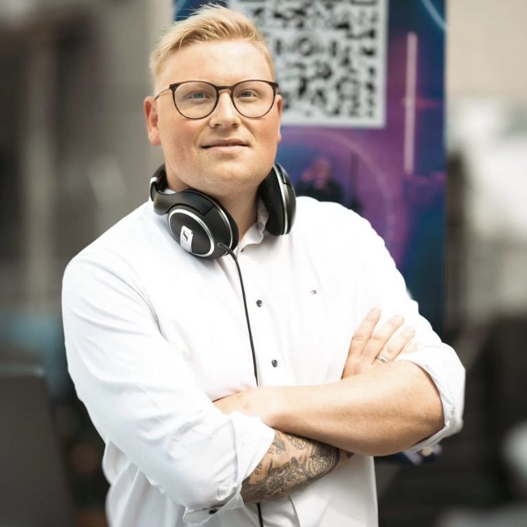 Ein Mann mit Brille und Kopfhörern lächelt, Arme verschränkt, vor moderner Hintergrundkulisse. DJ Rob spielt Schlager, Charts, 80er usw.