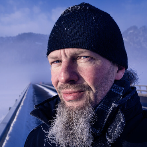 Mann mit grauem Bart und schwarzer Mütze vor einer verschneiten Berglandschaft.