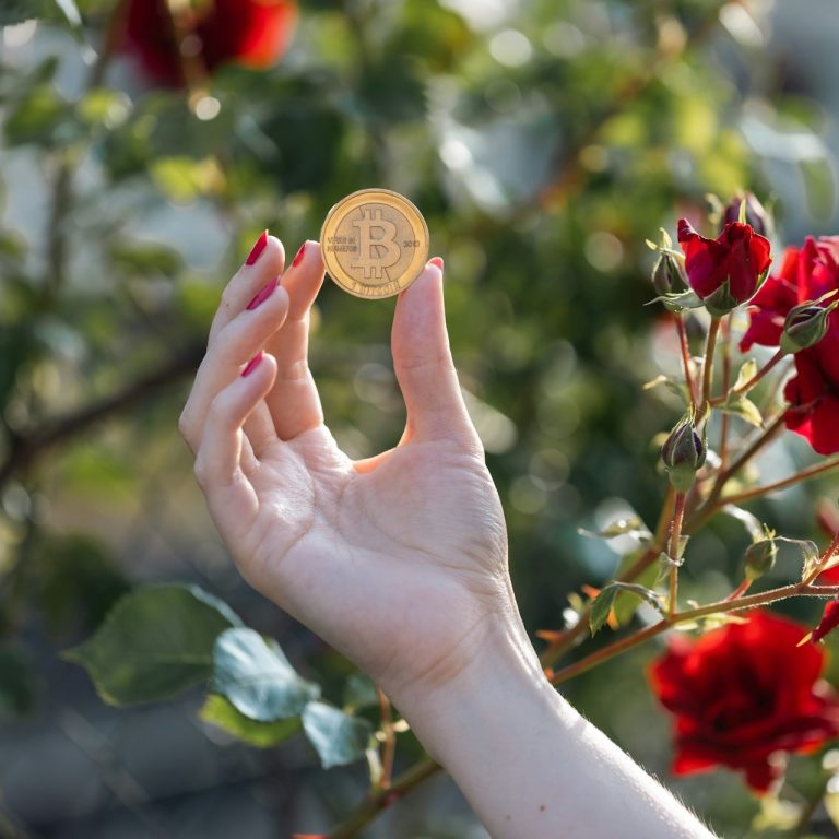 Eine Damenhand mit lackierten Nägeln hält eine goldene Bitcoin Münze vor roten Rosen.