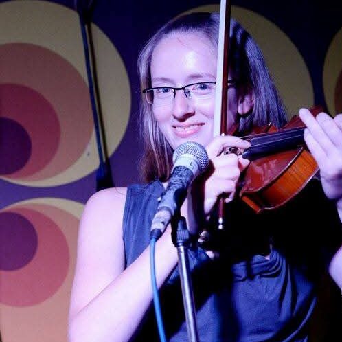 Junge Frau mit Brille spielt Geige und singt am Mikrofon auf einer Bühne. Bluenote Marta ist Violinistin/Geigerin.