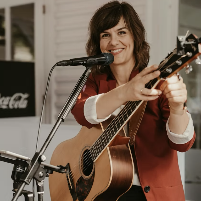 Frau mit Gitarre lächelt und spricht ins Mikrofon auf einer Bühne. Kate Rena Fleming ist eine Hochzeitssängerin aus Mönchengladbach.